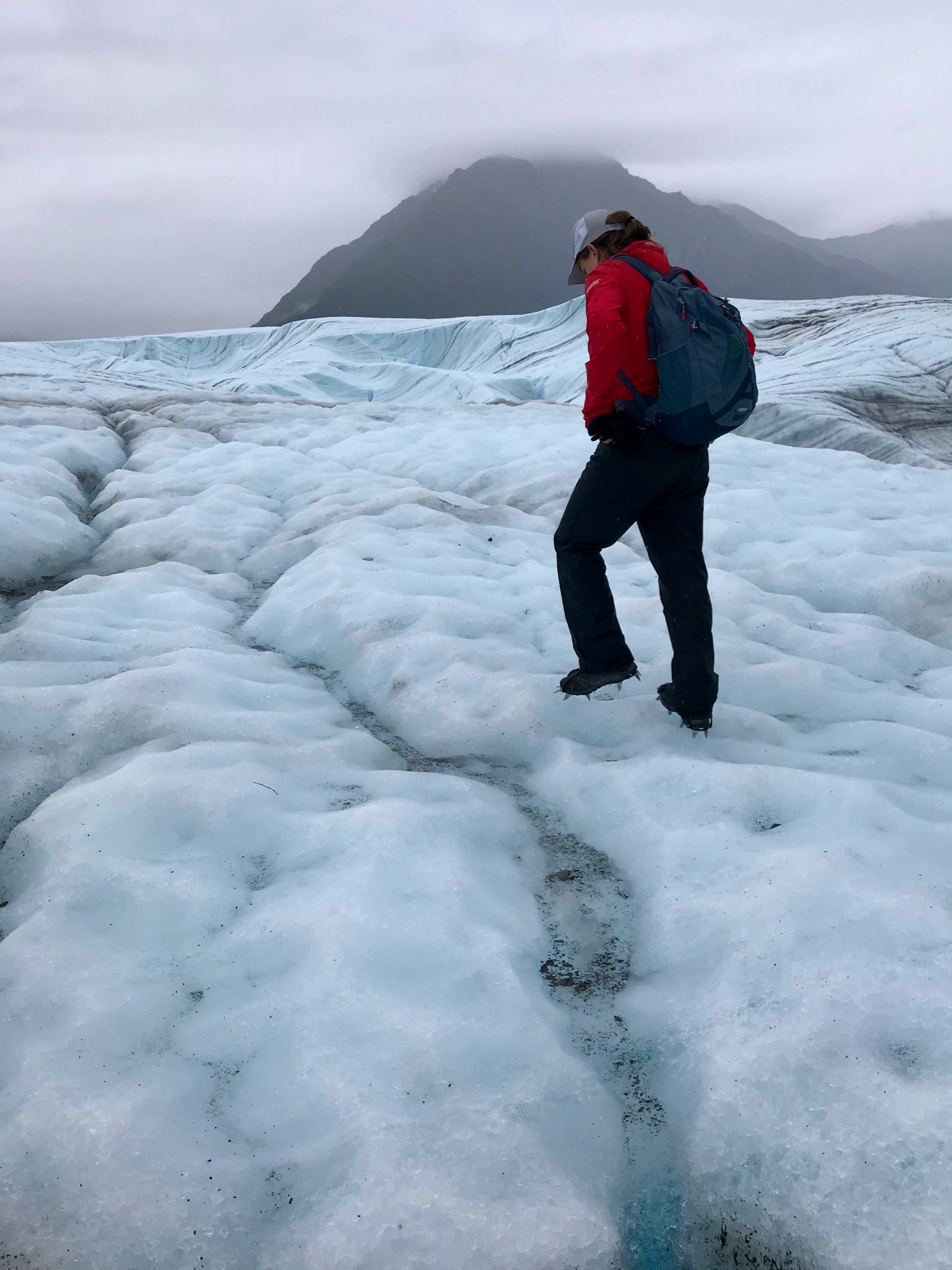 A woman wearing a red jacket, black pants, a backpack, and spiky shoes is seen from behind, walking along a glacier. There is ice surrounding her on the ground and a mountain in the distance. 