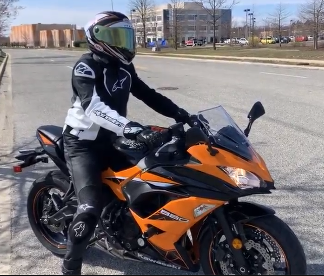 Person on a stationary orange motorcycle. The person is wearing a helmet, so you cannot see their face, and riding gear. Buildings at NASA Goddard are in the background. 