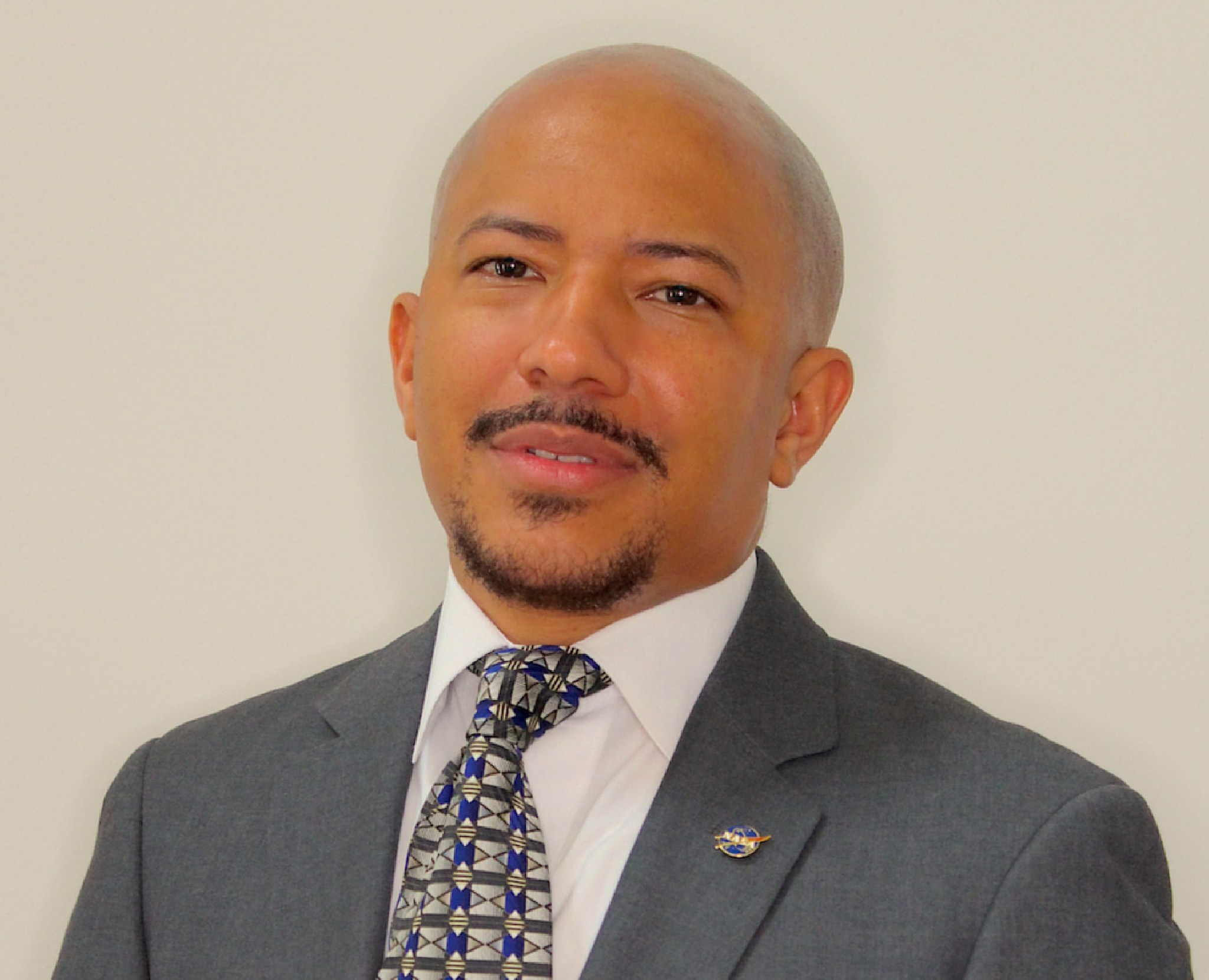 Man with tan skin and black facial hair, with a shaved head, wears a grey suit jacket, NASA pin, blue and grey patterned tie, and white dress shirt against a light tan background.