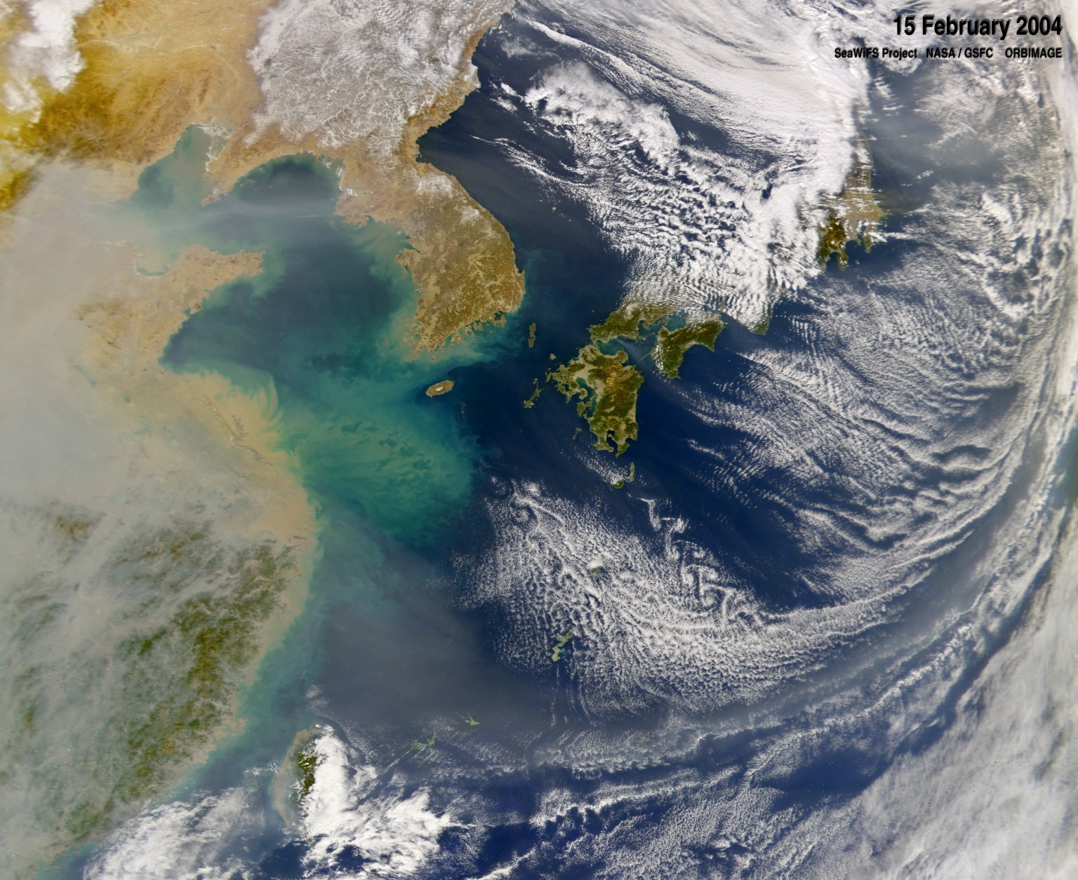 A satellite image of the Pacific ocean near eastern China, the Korean peninsula, and Japan. The land appears in shades of green and brown, with thick gray-brown clouds of pollution hanging over eastern China. The ocean is shades of deep blue and turquoise, and thin white clouds swirl over much of the right side of the image.