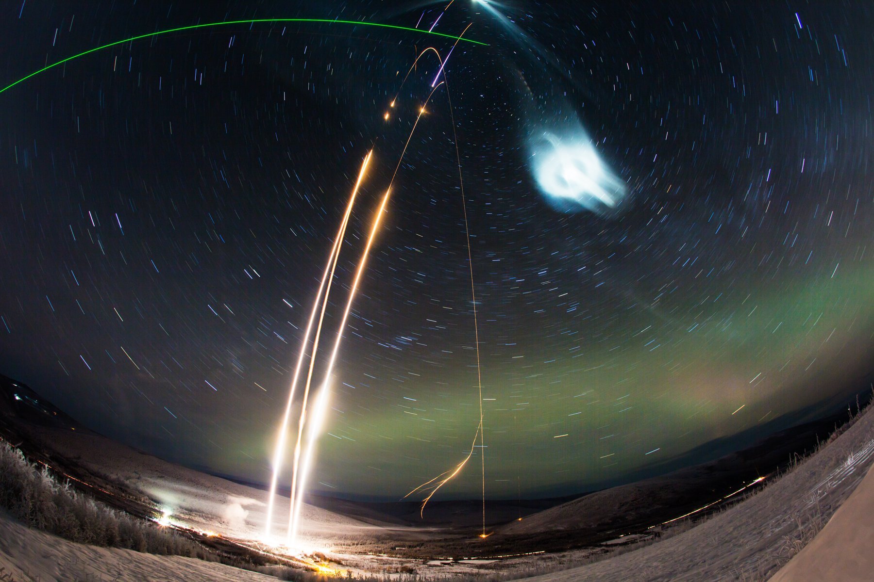 Three bright white streaks rise from the snow-covered ground on the left side of the image against a background of streaked star trails in the night sky. A roughly horizontal, green line curves along the top left of the image toward the top of the white streaks. A bluish-white cloud appears high in the sky to the right of the white streaks. The green glow of auroras appear in the lower right, near the horizon.