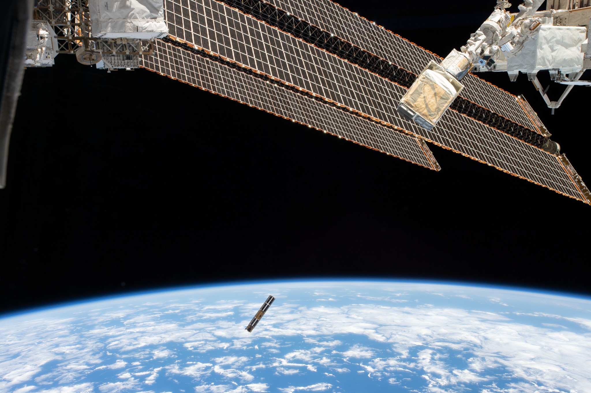 View of a long, narrow satellite in space, with the curve of Earth behind. Solar panels of the International Space Station are visible in the top half of the photo.