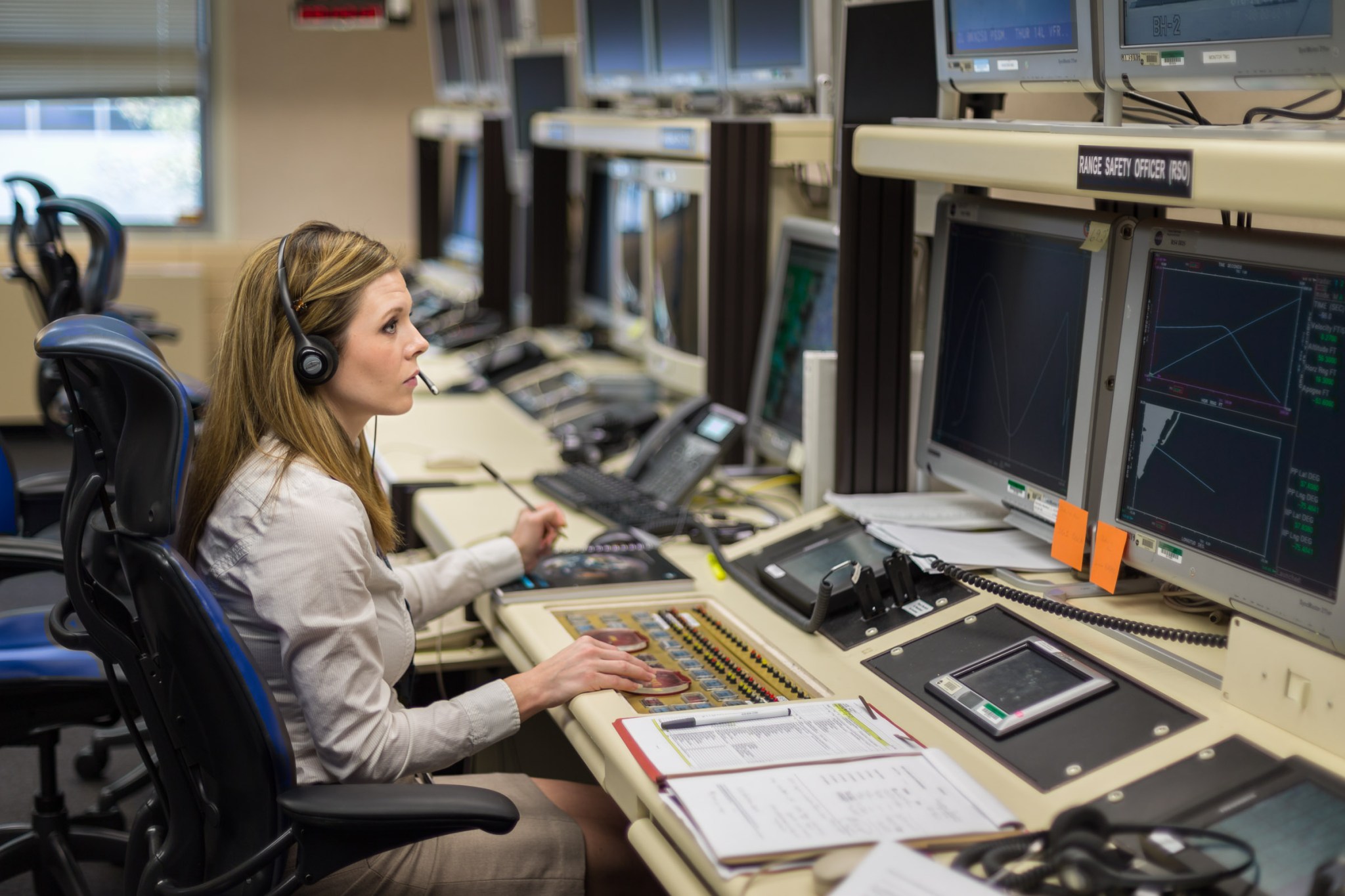 Woman with blonde hair wearing a white button down, tan skirt, and a headset  looks towards two monitors in a mission control room.