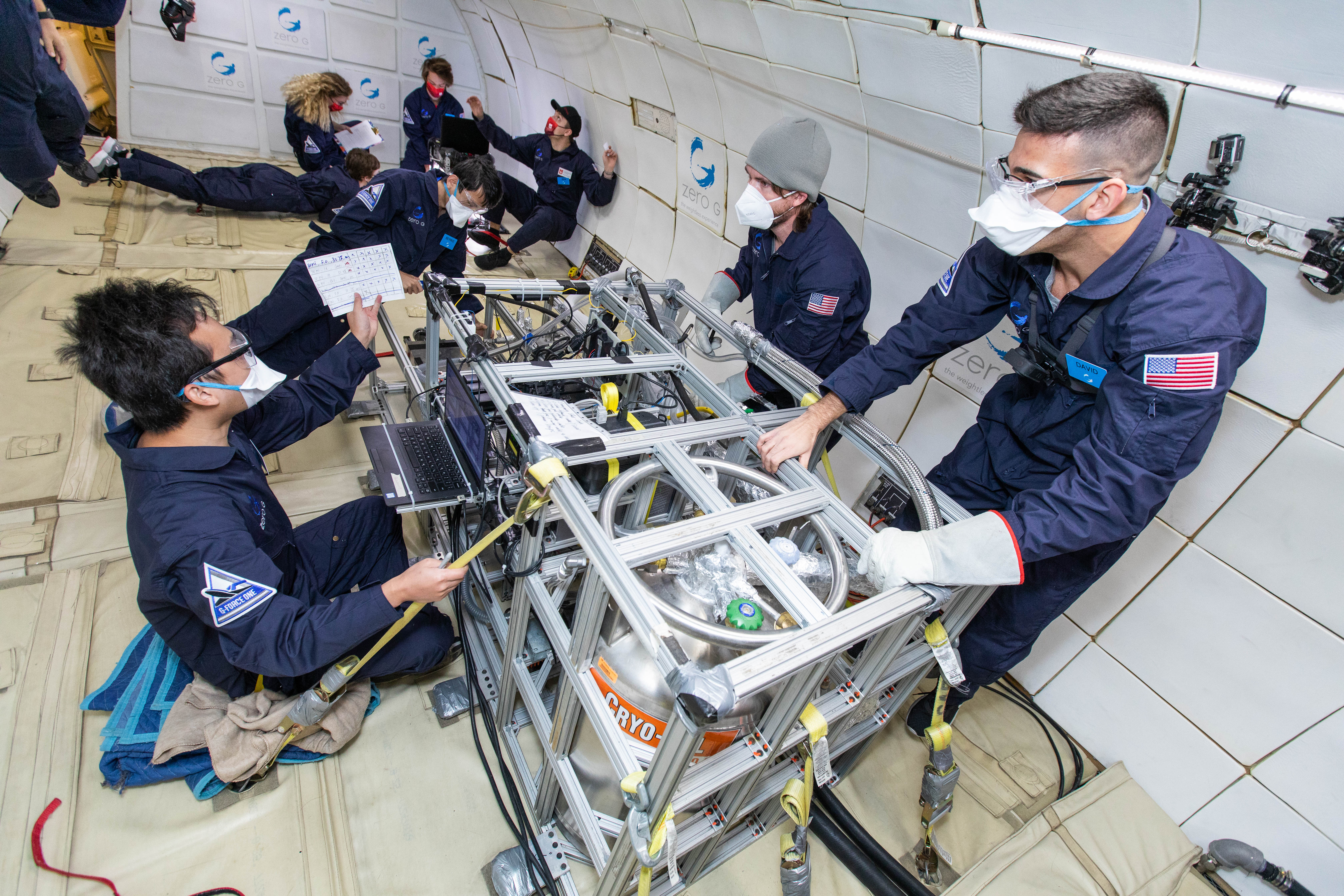 Researchers wearing masks monitor their experiment on a parabolic jet during microgravity. Other researchers are in the background.
