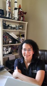 Image shows a woman posing for a photo at her desk. A bookshelf filled with space memorabilia is behind her.