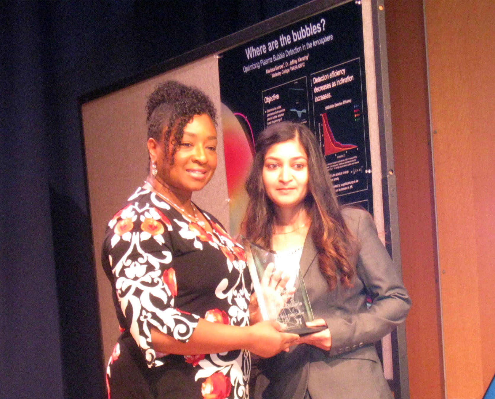 A woman with dark tan skin, brown, curled hair wearing a grey blazer holds an award that she's being presented by the woman standing next to her who has brown skin, black curly hair worn up, and is wearing a black, red, and white dress. They are standing in front of a poster that says "Where are the bubbles?"