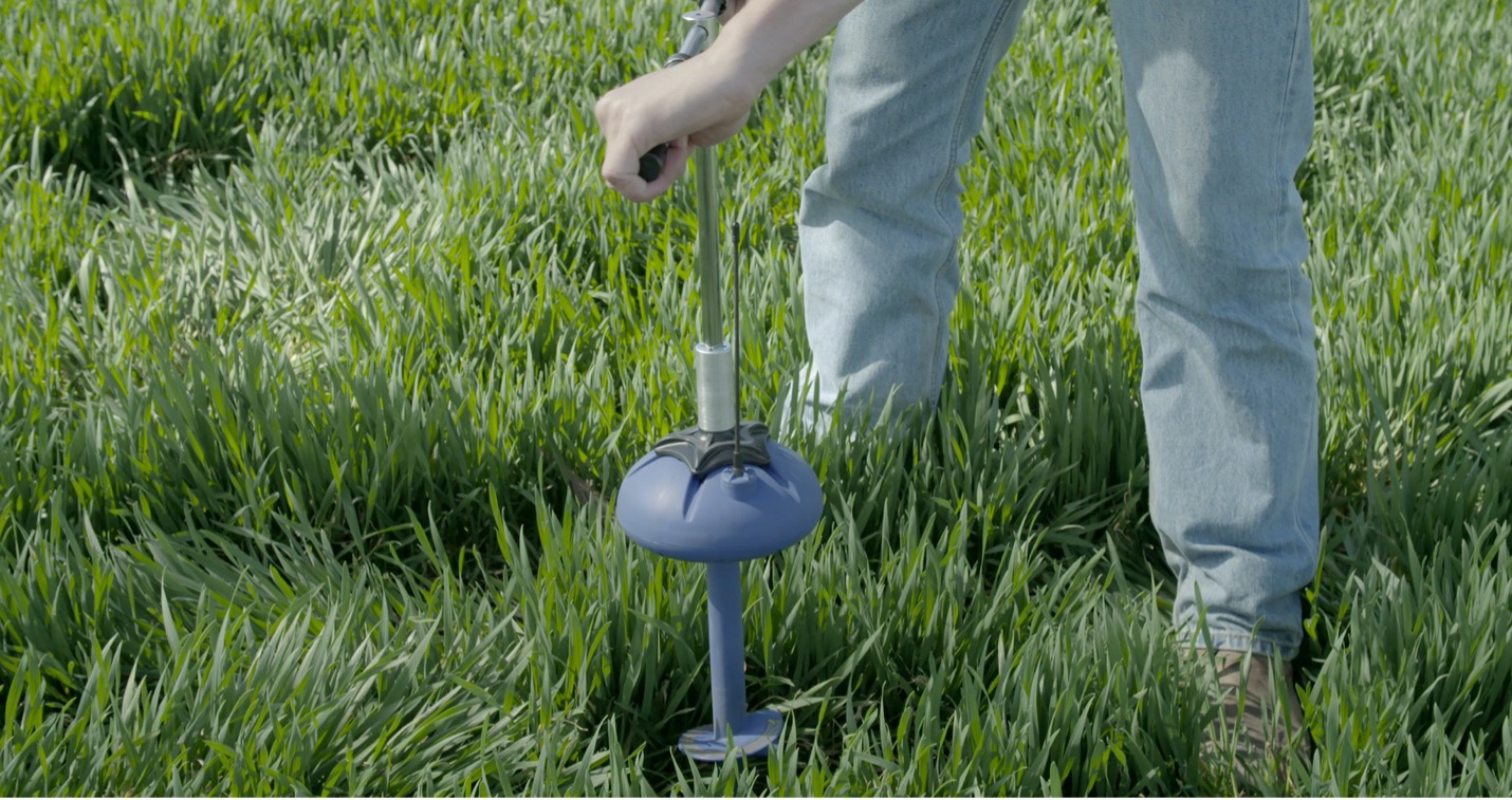 image of a shin-height soil quality sensor in lush green grass, the sensor looks like a blue mushroom, a person stands above with a tool, installing the sensor. The person is visible from the knee down.
