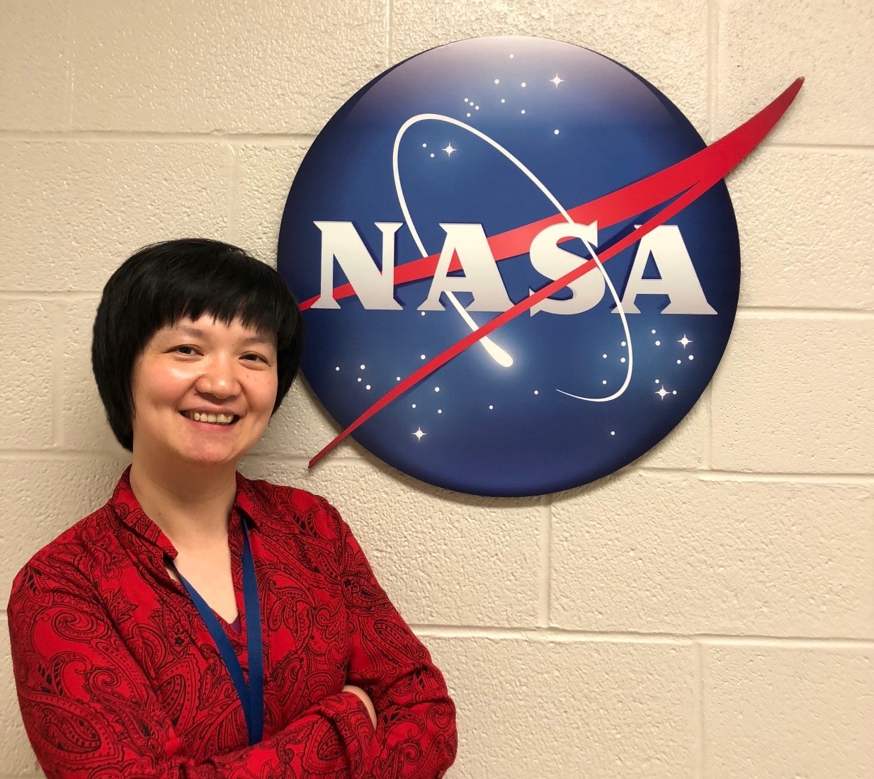 Woman with short, black hair, wearing a red shirt and a blue lanyard smiles in front of a wall with a large NASA meatball