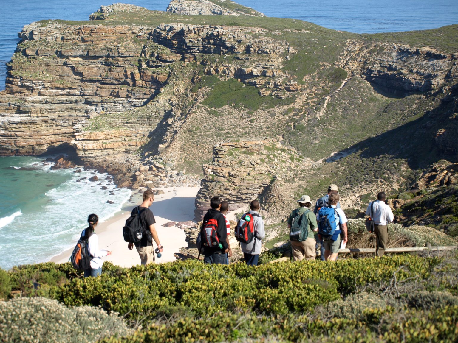 A group of students backpacks along a coastline in South Africa. The coast has steep, rocky cliffs partly covered with green grass and plants, extending down into clear turquoise water. Two of the cliffs encircle a small white beach. The students are of various genders and walk along a ridge in the foreground.