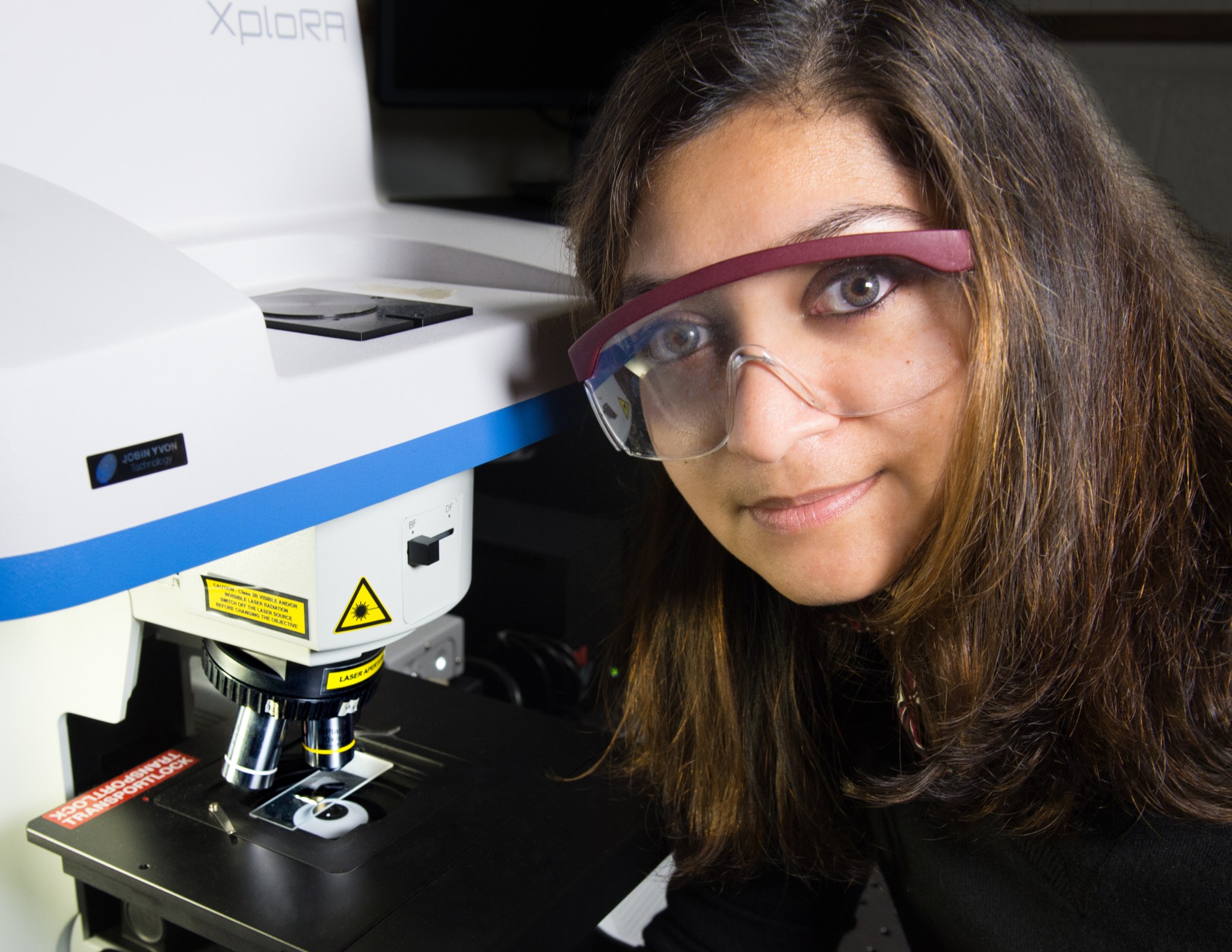 A woman with light brown skin and long dark brown hair wears burgundy-rimmed safety glasses, a black top and blue rubber gloves, leaning forward in front of a large microscope. The gray-white microscope takes up most of the left side of the image, with a blue stripe along its middle edge and the logo "XploRA" at the top. Underneath the lens in use is a slide resting on a platform, and a red label reading "Transport Lock" at the edge of the platform.