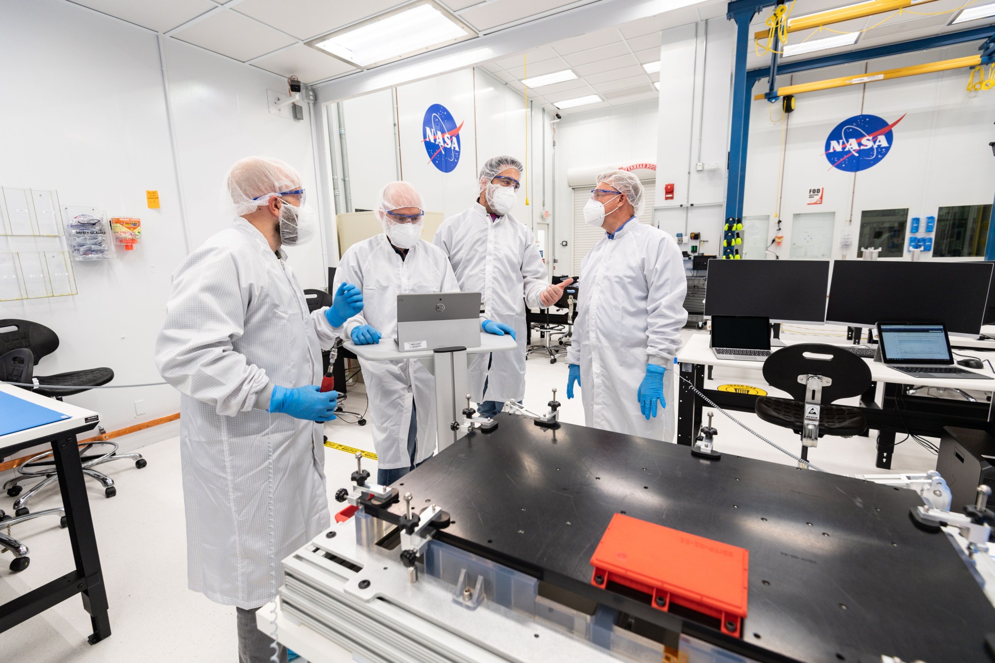 Four people wearing hairnets, goggles, face masks, gowns and blue gloves stand behind a flat, black piece of hardware in front