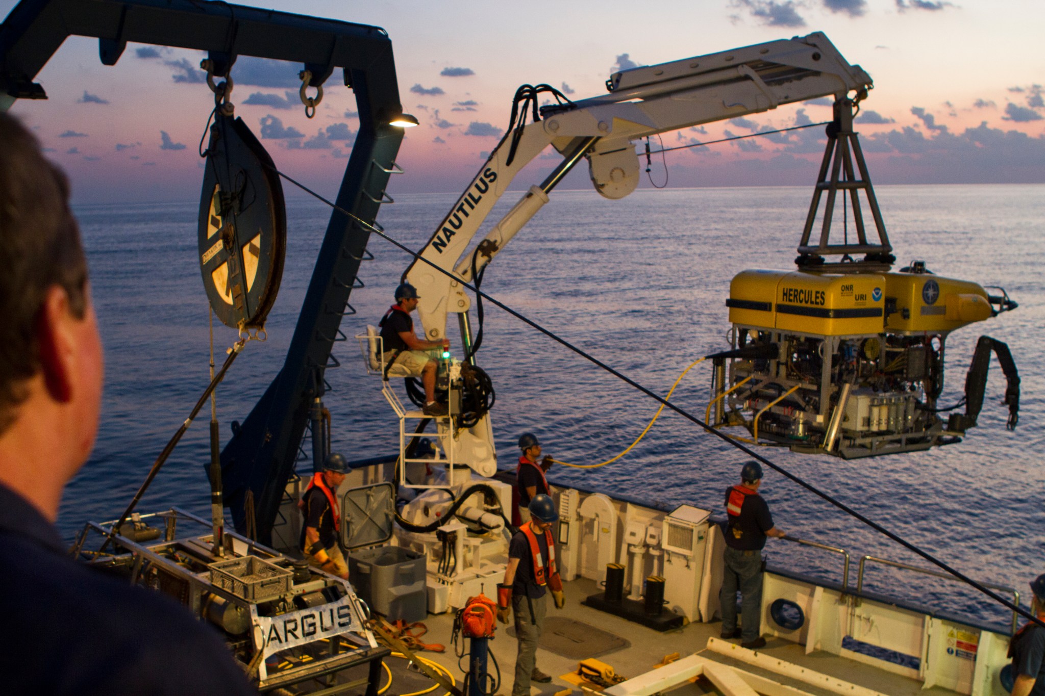 Crane holding up a robotic explorer from a boat at sea.