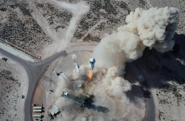Aerial view of the New Shepard vehicle above the launch pad, with dust billowing.