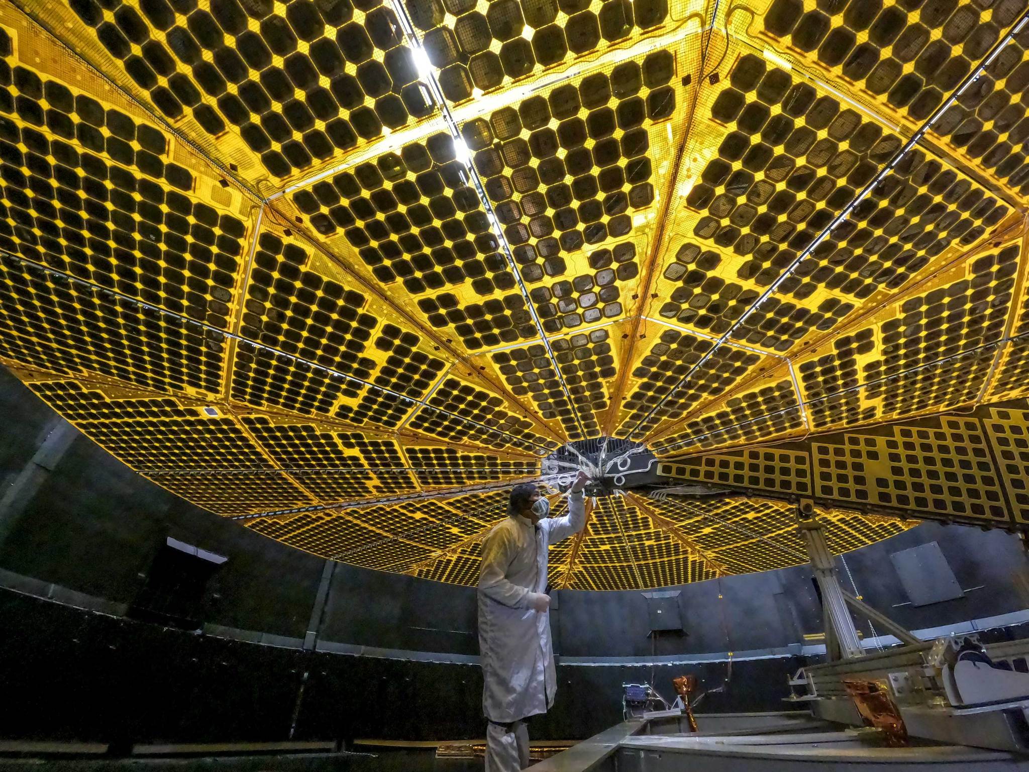 Lucy's solar panels extended to form a flat circle of yellowish panel, with small gold circles covering the surface. The panel is open high in the air, like an umbrella. A person in a clean room stands below the