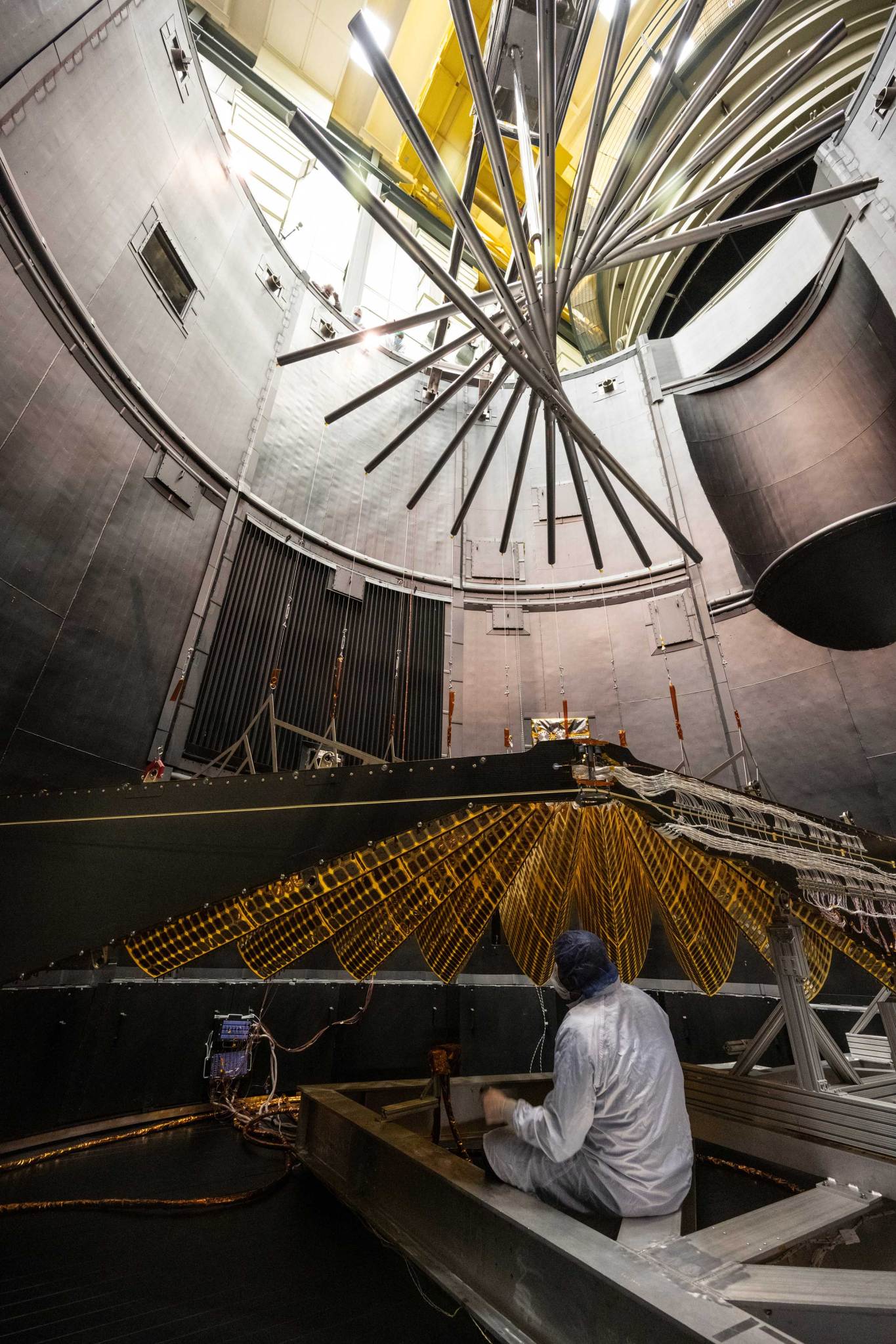 Inside of an industrial-gray testing chamber with a spiral of support beams above a folded, gold-hued solar panel. A person in a white clean room suit is seated on a beam looking up at the panel.