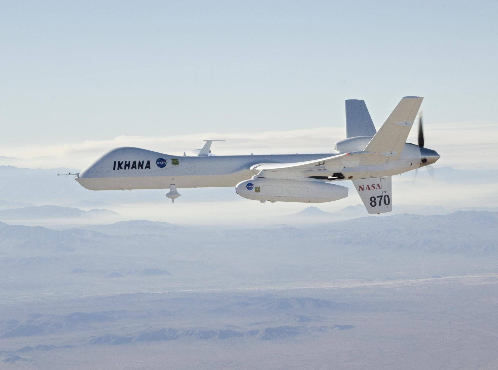 A large drone aircraft flies high above a mountainous area,