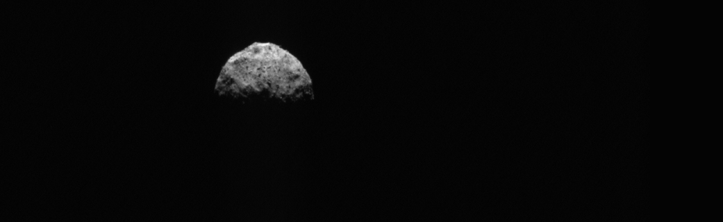 A ghostly gray image of asteroid Bennu against black backdrop of space. The asteroid is visible in gibbous form, with the bottom half swallowed by space.