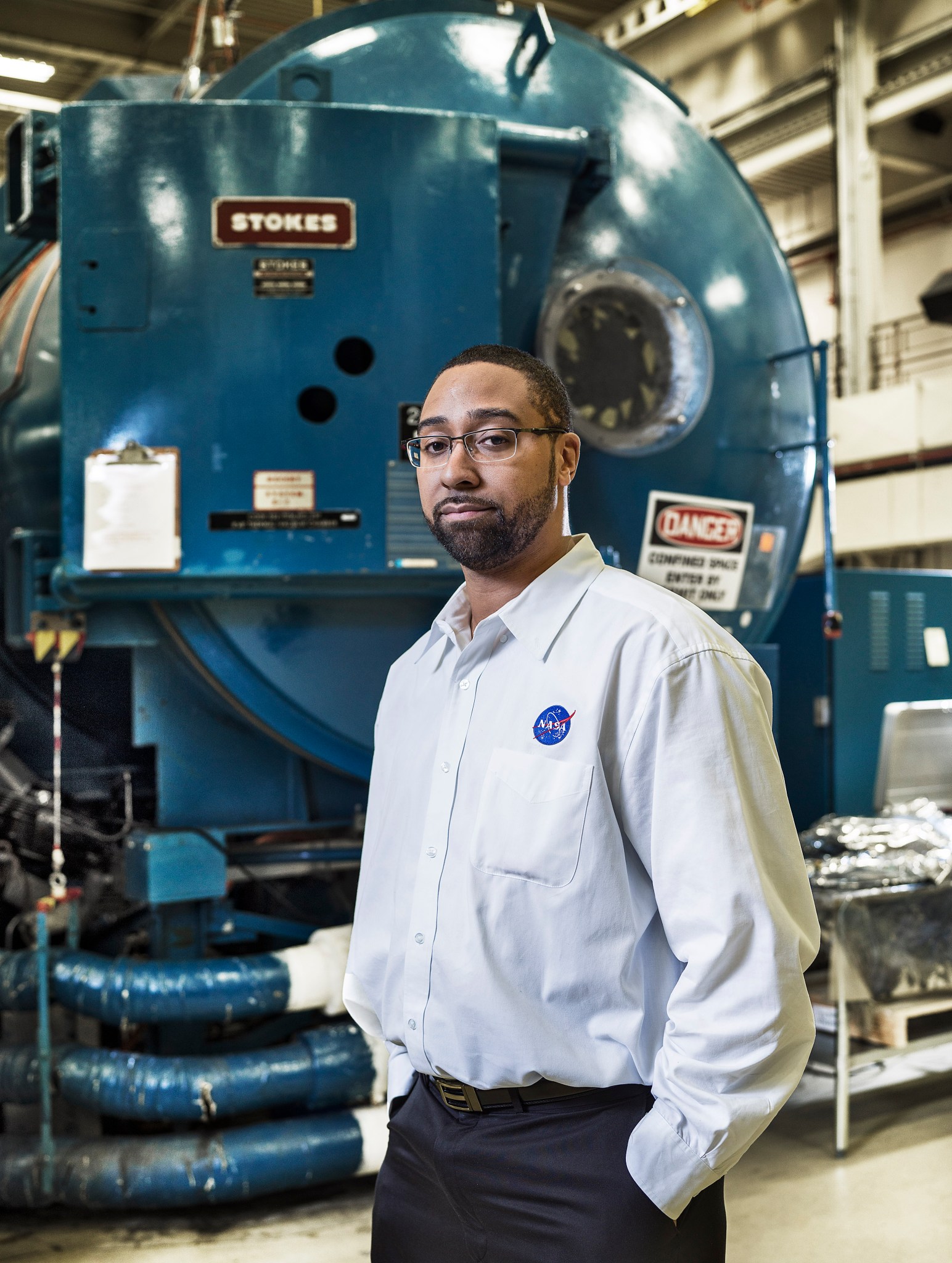 A Black man with short black hair, wearing glasses, a white dress shirt with a NASA logo, and navy dress pants stands in front of a large blue machine in the environmental testing facility at NASA Goddard.   