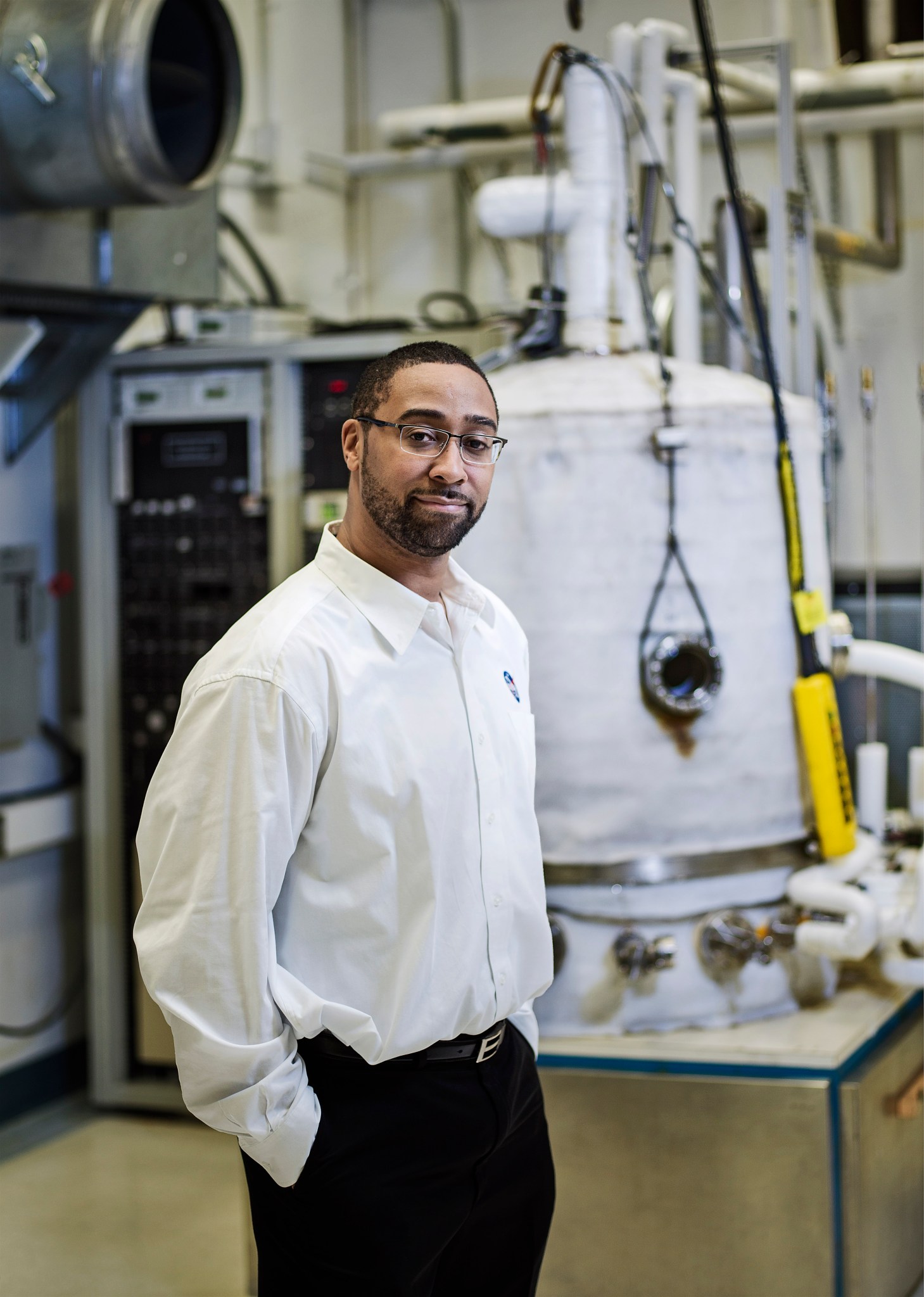 A Black man with short black hair, wearing glasses, a white dress shirt with a NASA logo, and navy dress pants stands in front of machines in the thermal engineering branch lab at NASA Goddard. 