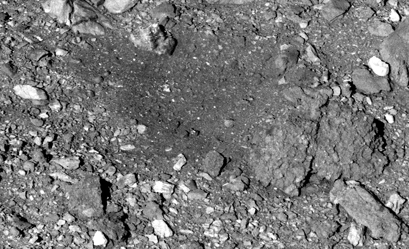 A black and white image a rocky rubble pile where OSIRIS-REx captured a Bennu sample. Most of the rocks are roughly the same gray color as the background, but a few are a brighter white. In the center of the image, the rocks appear slightly darker.