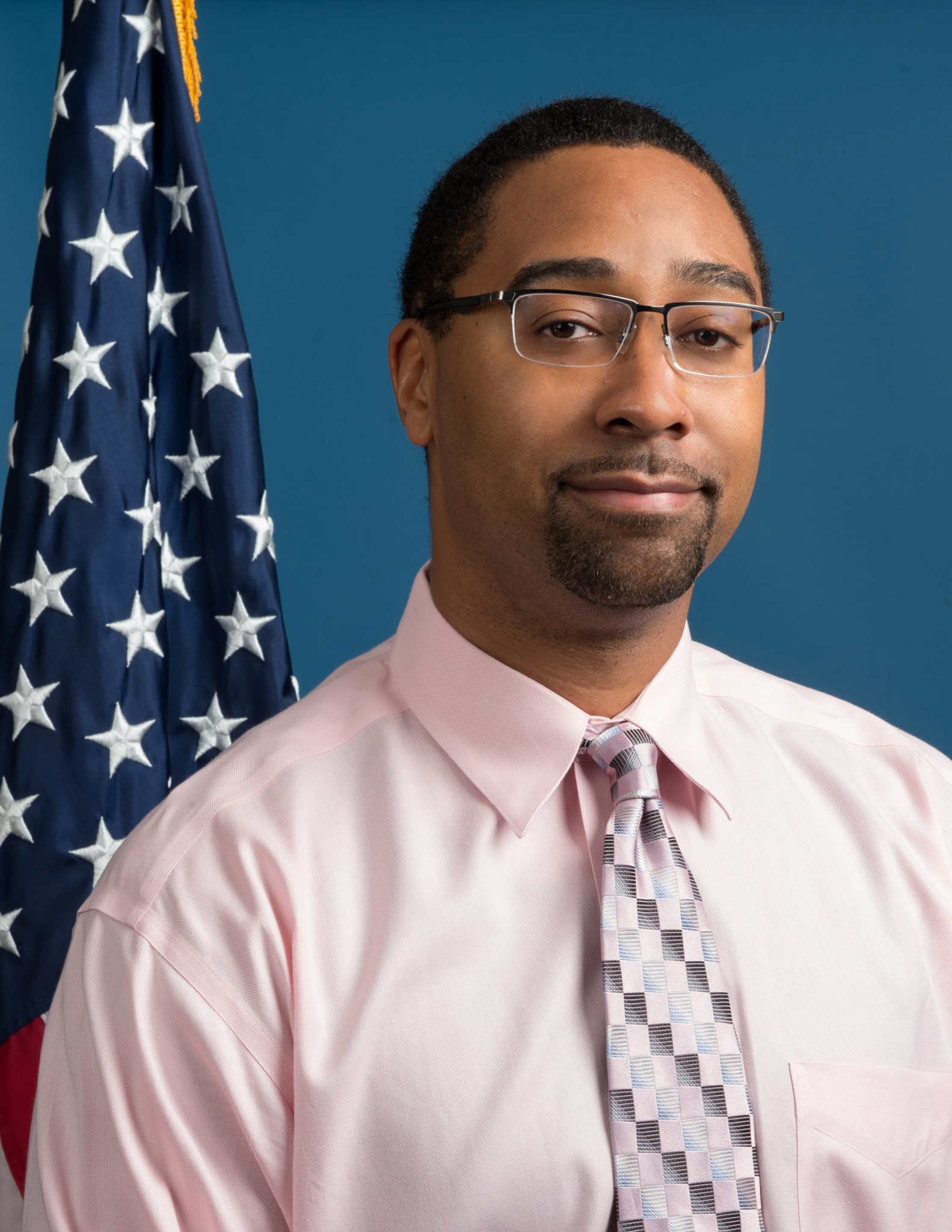 A Black man with short black hair wears glasses, a pink shirt, and pink and grey tie. He smiles against a blue background with the American flag on the left. 