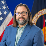 Headshot of a white man with shoulder-length straight brown hair, a brown and grey beard, and glasses with black rims. He is wearing a light blue buttonup and a royal blue suit.