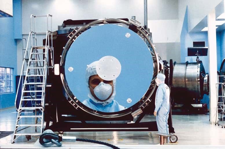 Old image of a scientist in a clean room suit looking into Hubble's primary mirror and it reflecting much larger in the mirror