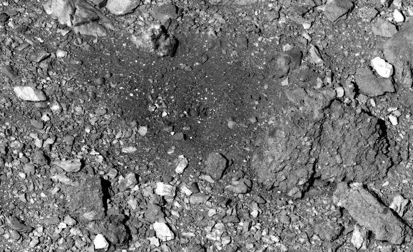 A black and white image a rocky rubble pile where OSIRIS-REx captured a Bennu sample. Most of the rocks are roughly the same gray color as the background, but a few are a brighter white. In the center of the image, the rocks appear darker.