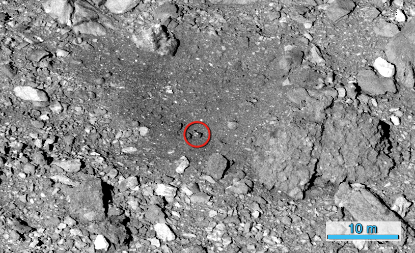 Gif of the gray, pebbly surface of asteroid Bennu. In the first image, a red circle around a rock near the center moves to the same rock much lower in the image in the second frame. A small red X appears over the center of the image in the second frame.