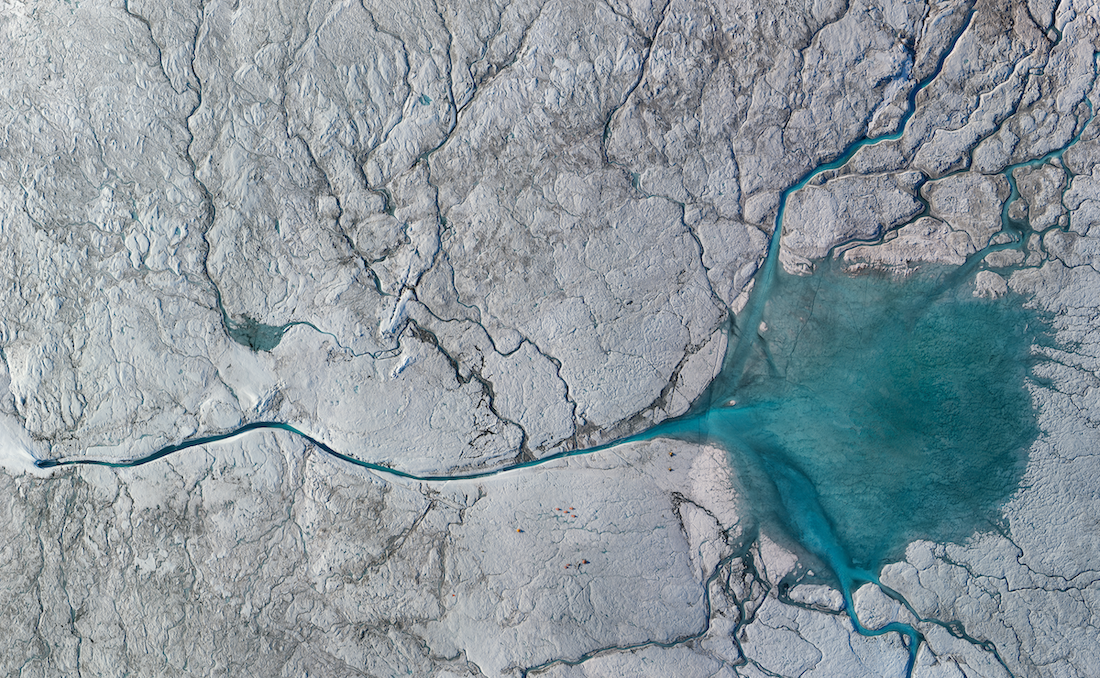 An aerial photo of a glacial river in Greenland. The river looks like a thin turquoise line emerging from the snow-covered ice sheet and flows across the image to a large, fan-like pool of turquoise water near the right side. Cracks and channels in the ice spread out from the river, shaded gray.