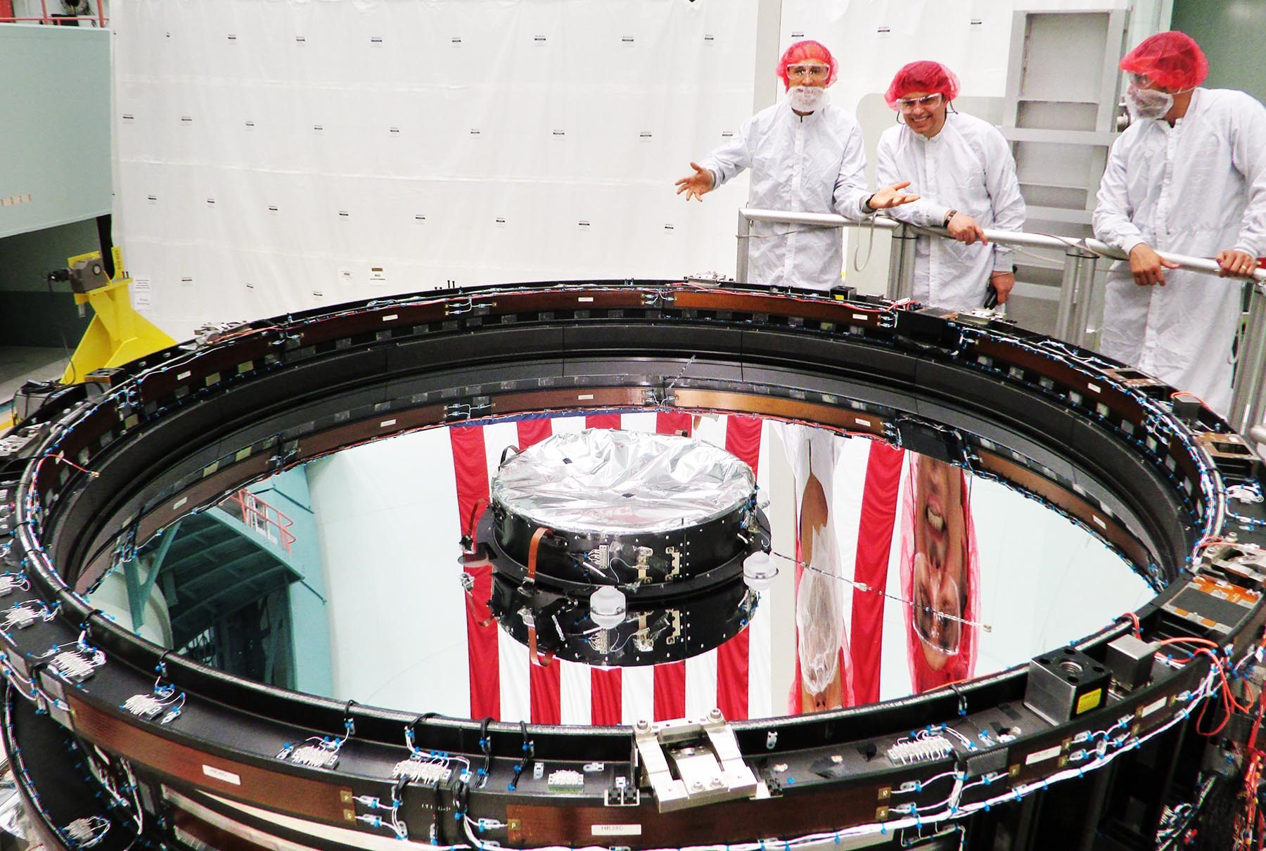Three men wearing white lab coasts and red hair nets stand over a large round mirror