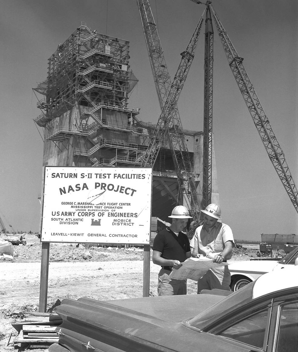 Two men review plans in the foreground with the A-2 test stand under construction in the background. A sign in the photo reads Saturn S-II Test Facility, NASA PROJECT
