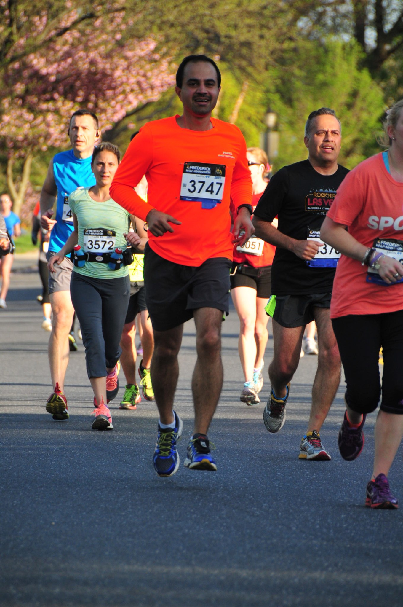 Man with tan skin and black hair wears an orange long sleeve shirt and black shorts and is running in a race. There are many other runners around him. 