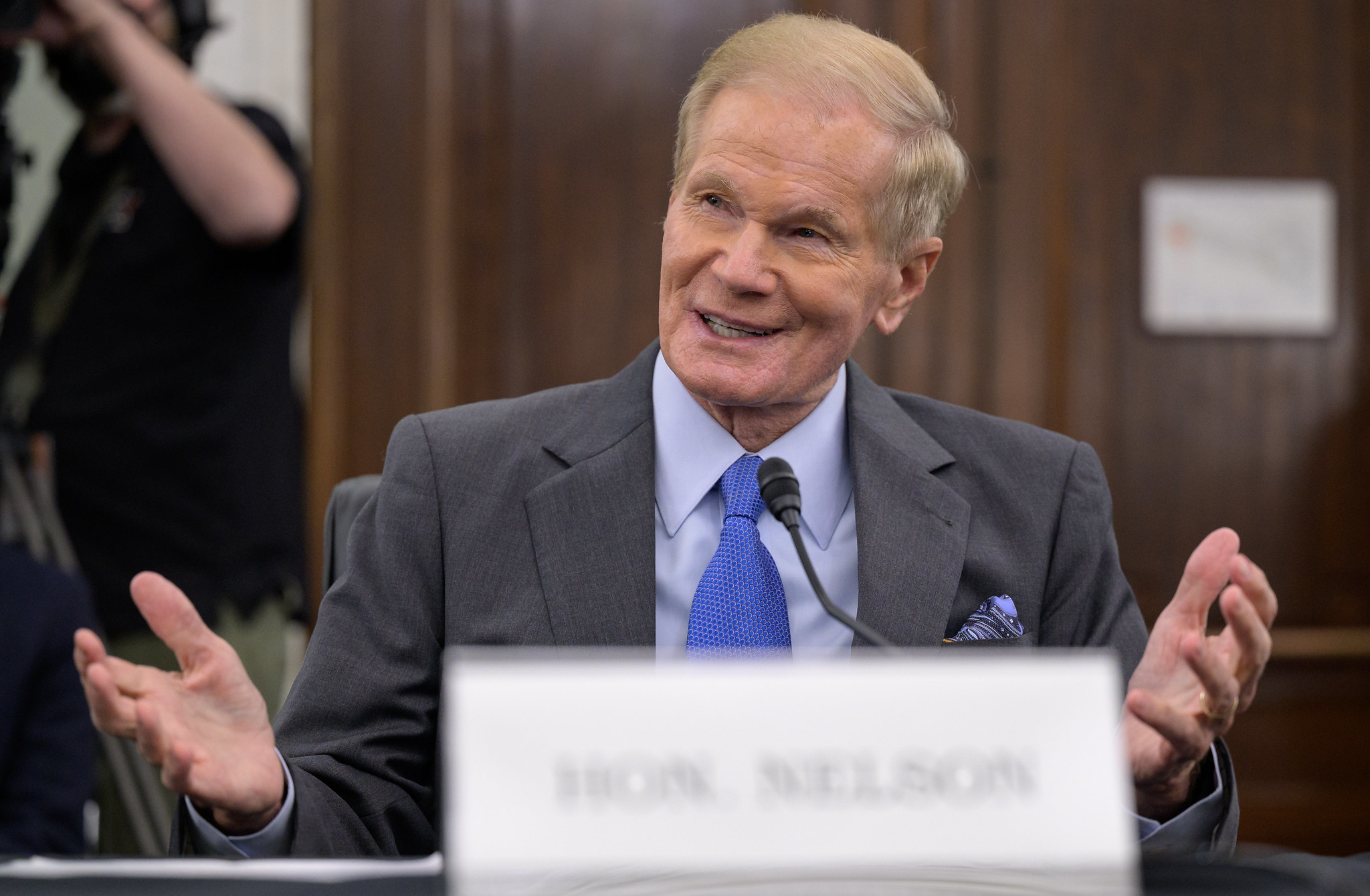 This image shows Bill Nelson, former U.S. senator and confirmed NASA administrator, smiling with his hands open while seated at the table before the Senate Committee on Commerce, Science, and Transportation. He is wearing a gray suit, blue shirt and tie.