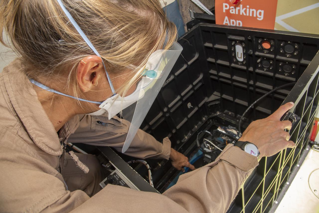 A woman with blonde hair pulled back in a ponytail leans over a crate containing a science camera system, performing pre-flight checks. She wears brown coveralls, a white mask covering her nose and mouth, and a clear face shield. The camera looks like a collection of wires and metal in the bottom of a black crate.
