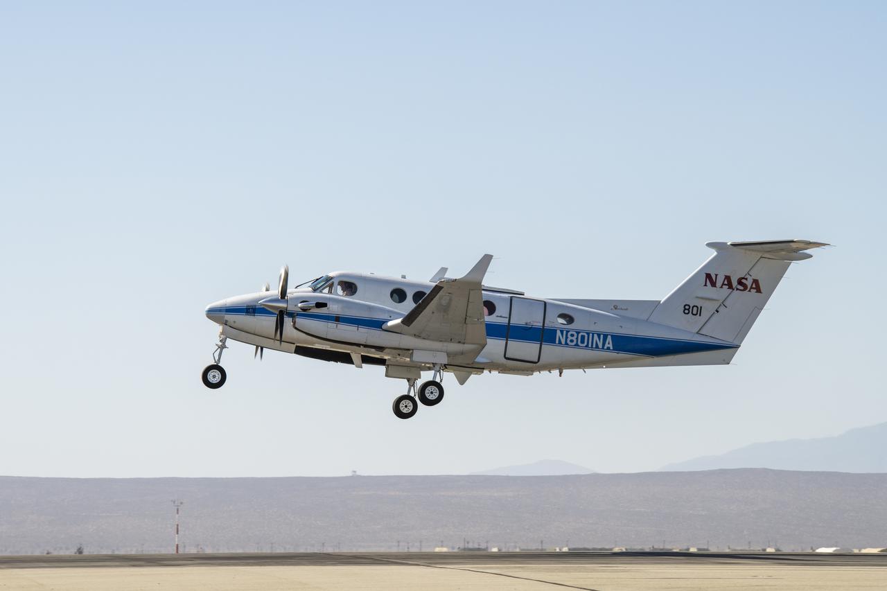 A photograph of a small twin-engine airplane taking off. The plane is silver-white with a blue stripe running from nose to tail, and has twin propellers. The NASA logo is visible on the tail. Mountains and gray-brown trees are visible in the far distance.