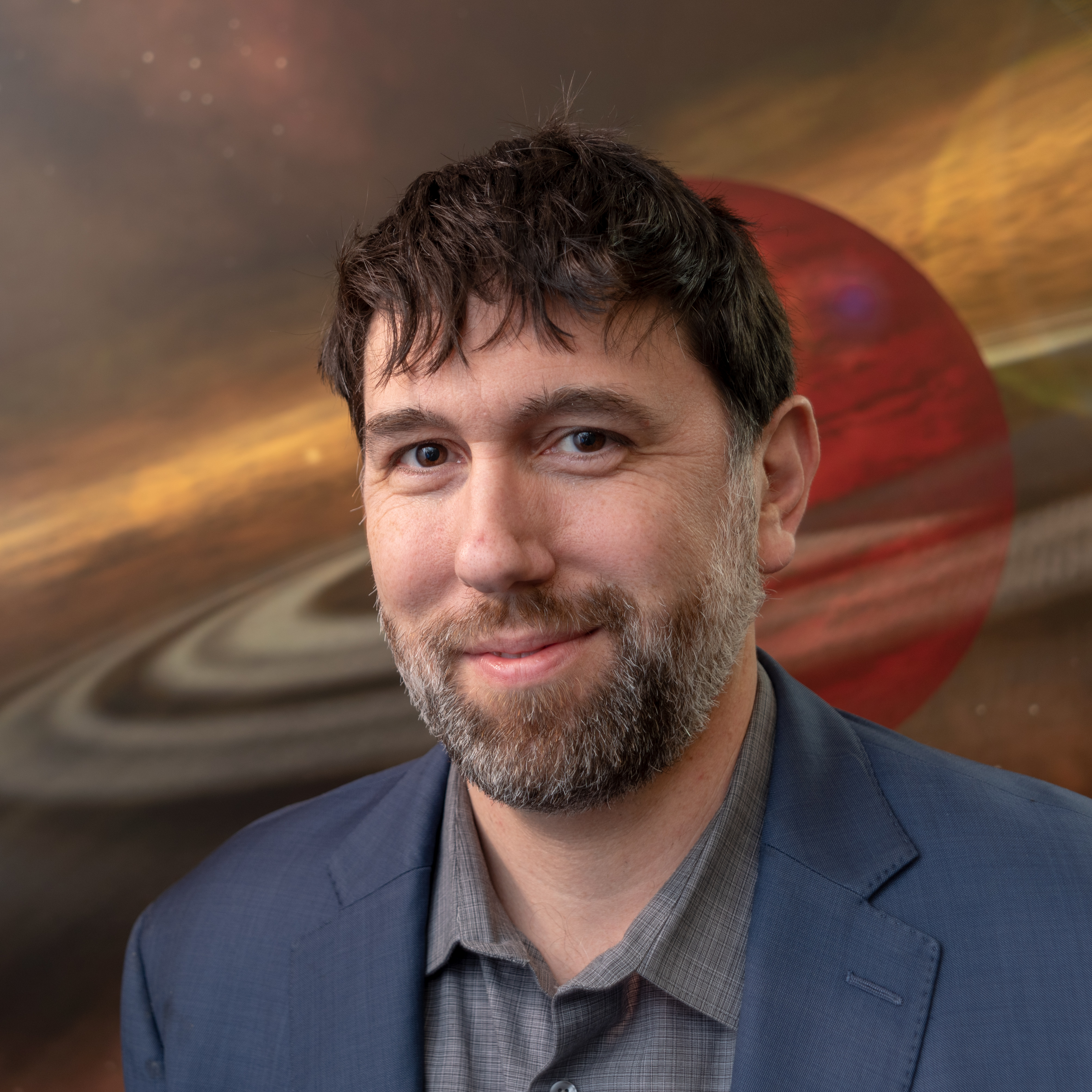 Man fair skin, brown hair and brown and grey facial hair wears a grey shirt and blue blazer. His headshot is in front of an illustration of Saturn in shades of red, yellow and brown.