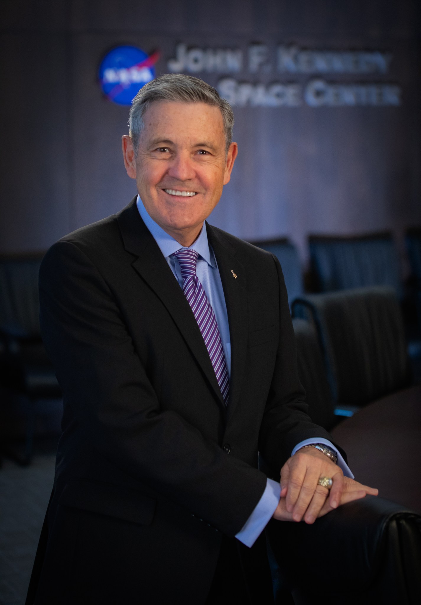 Portrait of Bob Cabana smiling in a black suit, his hands resting on a meeting room chair.