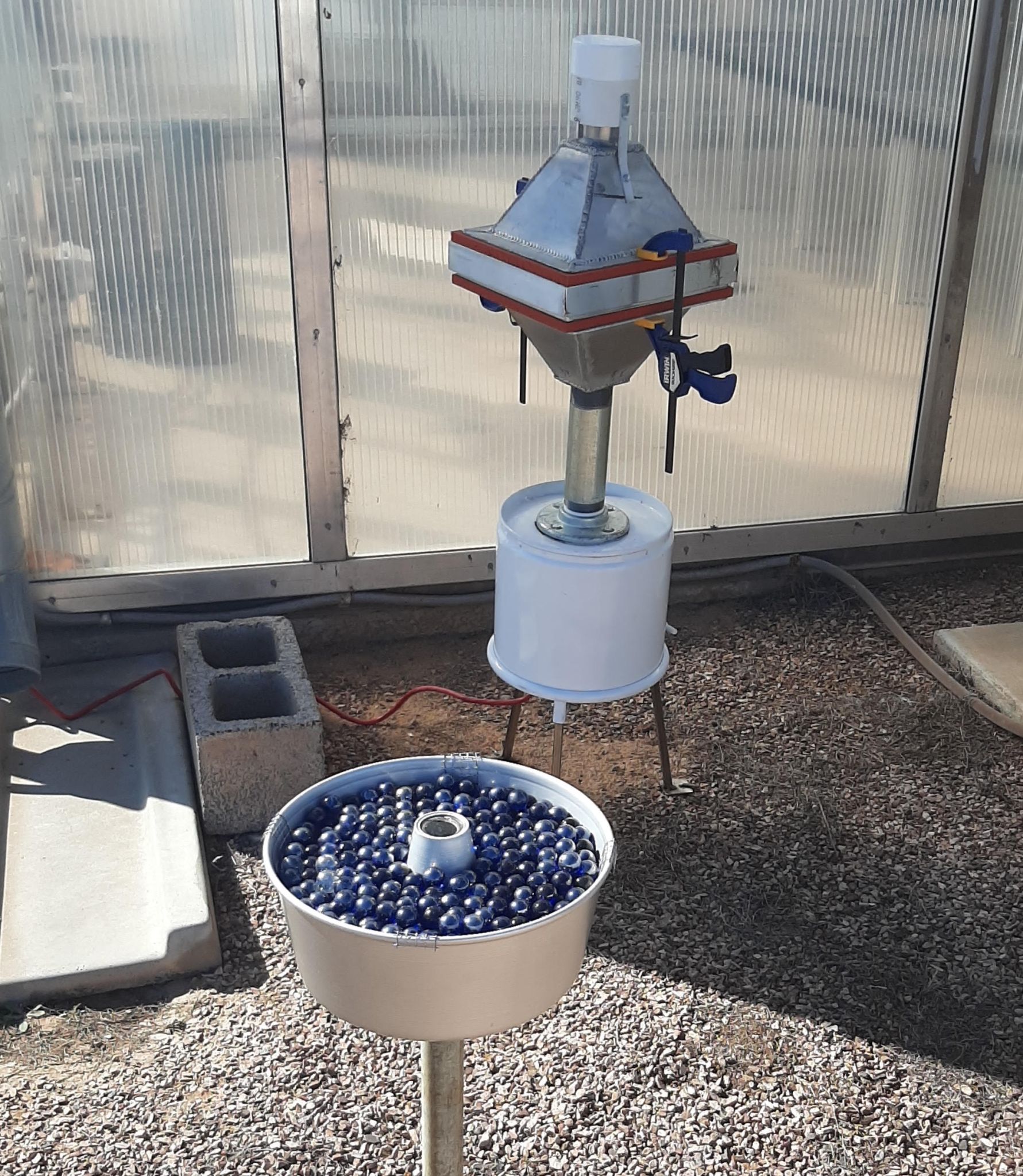 A bundt cake pan filled with blue marbles sits on a tall thin pip in front of a second instrument made of two pyramids stacked with bases together.