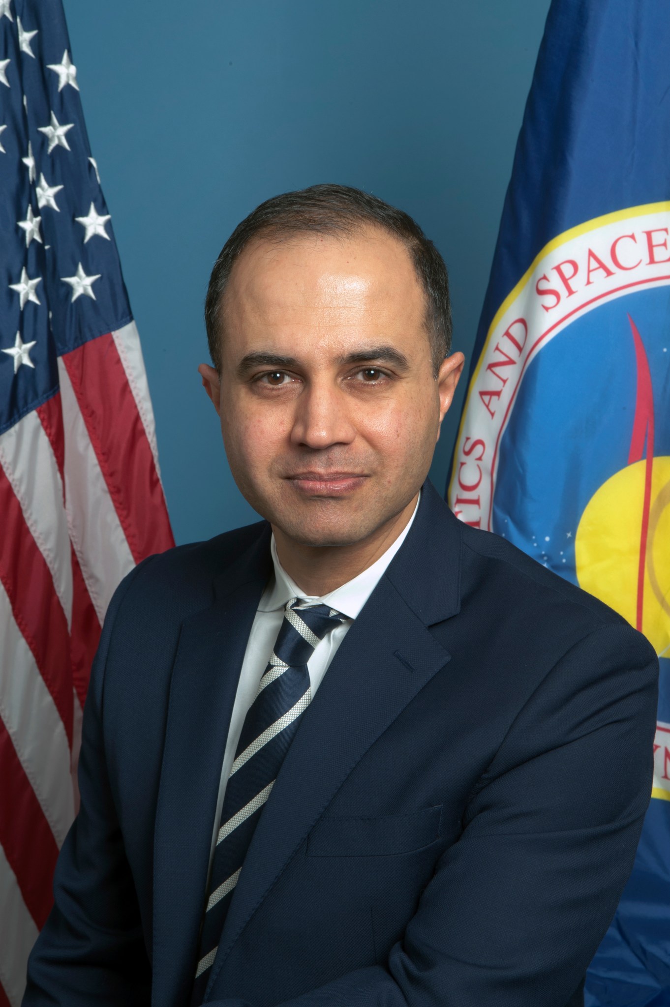 Man with tan skin and black hair wears a navy suit with a navy and white tie against a blue background with the American flag on the left and the NASA flag on the right. 