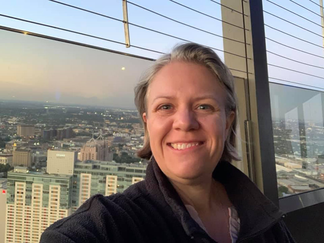 A blonde, fair skinned woman smiles while taking a selfie in front of windows overlooking the cityscape of San Antonio.