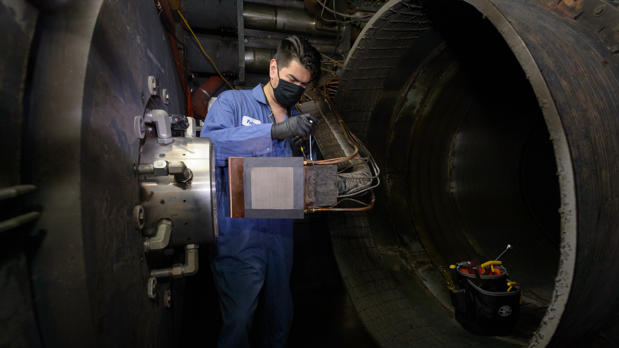 A man in a blue jumpsuit and black gloves and mask installs an object on a metal arm in front of the opening to a large tunnel on the right in which a toolbox sits.
