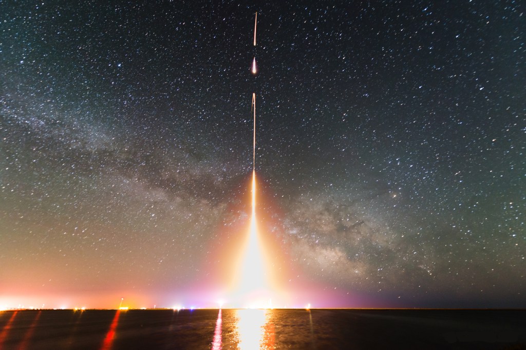 A bright, yellow, vertical rocket trail rises from a dark, watery horizon and into a starry night sky with the Milky Way extending across the background. At the bottom, near the horizon, the rocket trail fans out into a bright, triangular glow, wider at the bottom and narrowing farther upward. Near the top of the view, the trail breaks up with two gaps separating parts of the trail.