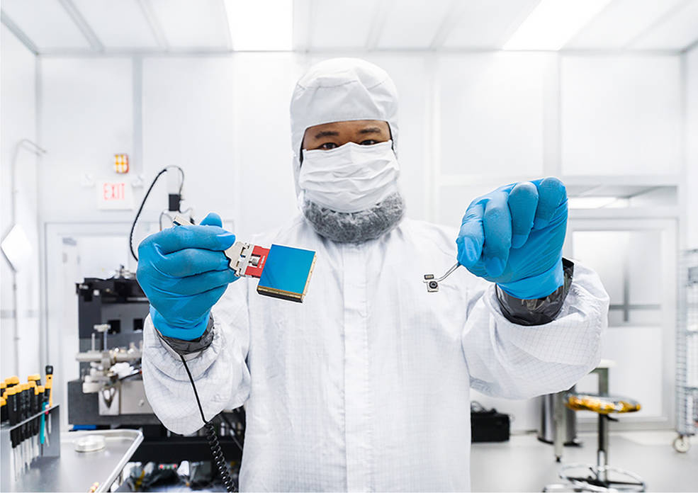 A man with brown skin is dressed in a white lab coat, white hat, white face mask, and blue gloves. He holds a Saltine-sized blue detector in one hand and a small cell phone camera with tweezers in the other hand.