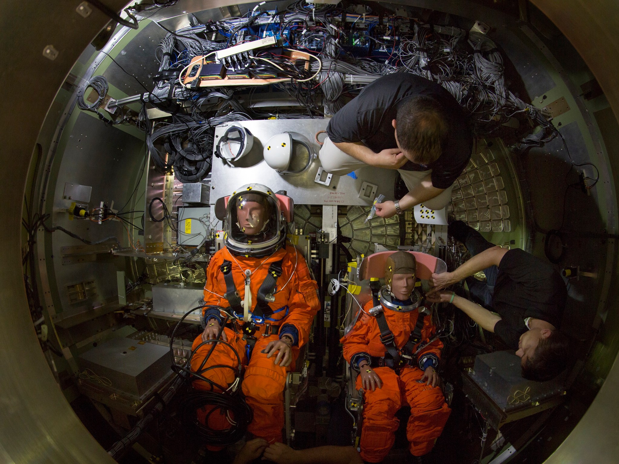 Two dummies in orange spacesuits are situated in chairs in a mockup.