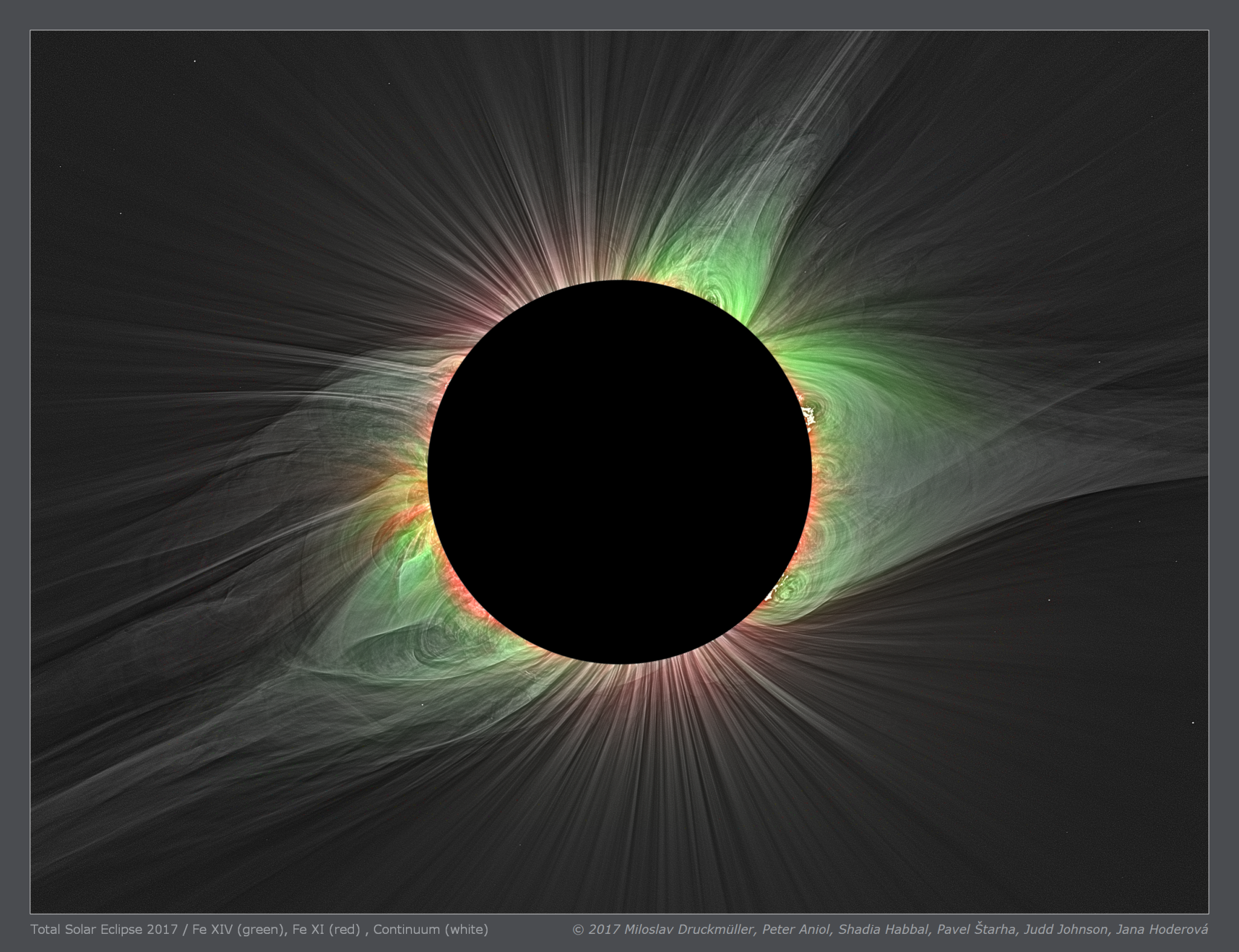 An eclipse image shows the Sun's face covered by the Moon, revealing the corona. The area above the Sun's north and south poles are streaked with red. On either side, the petal-shaped streamers of the corona are mostly green, with some red close to the solar surface. 