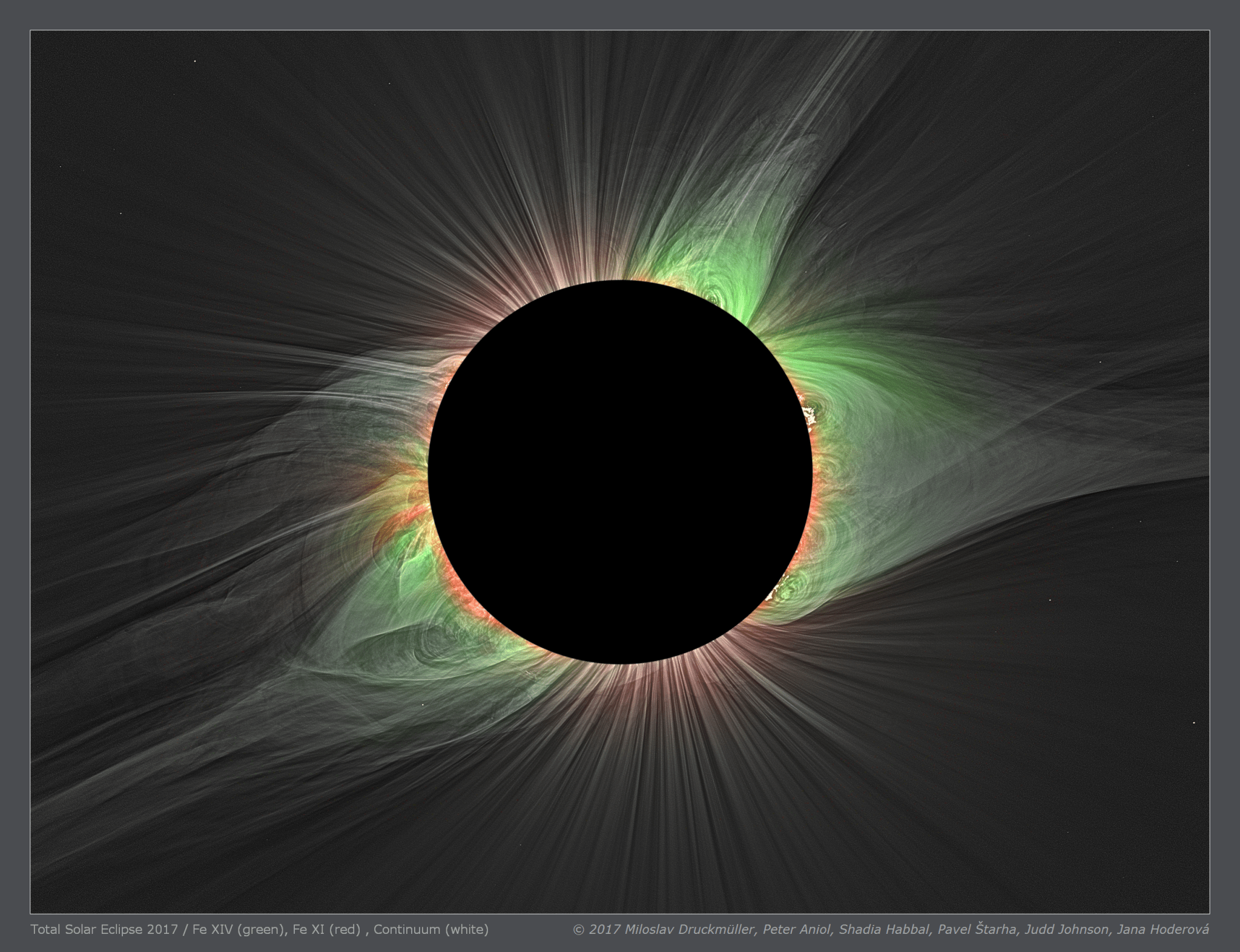 An eclipse image shows the Sun's face covered by the Moon, revealing the corona. The area above the Sun's north and south poles are streaked with red. On either side, the petal-shaped streamers of the corona are mostly green, with some red close to the solar surface. 