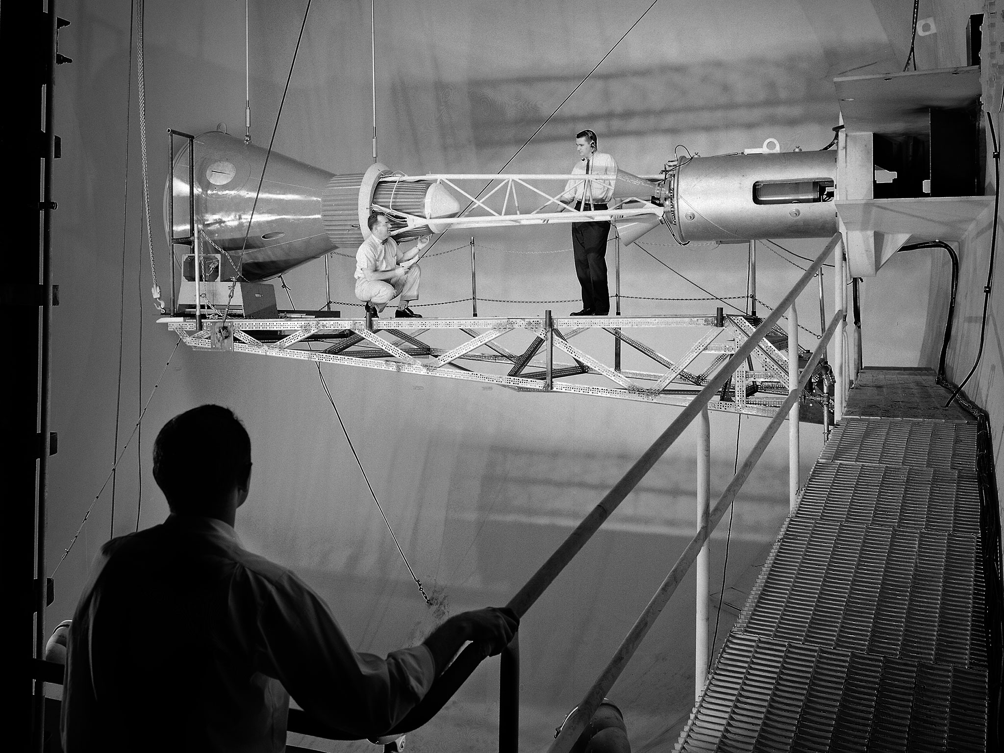 A rocket engine inside a test facility with two engineers walking around it on scaffolding.