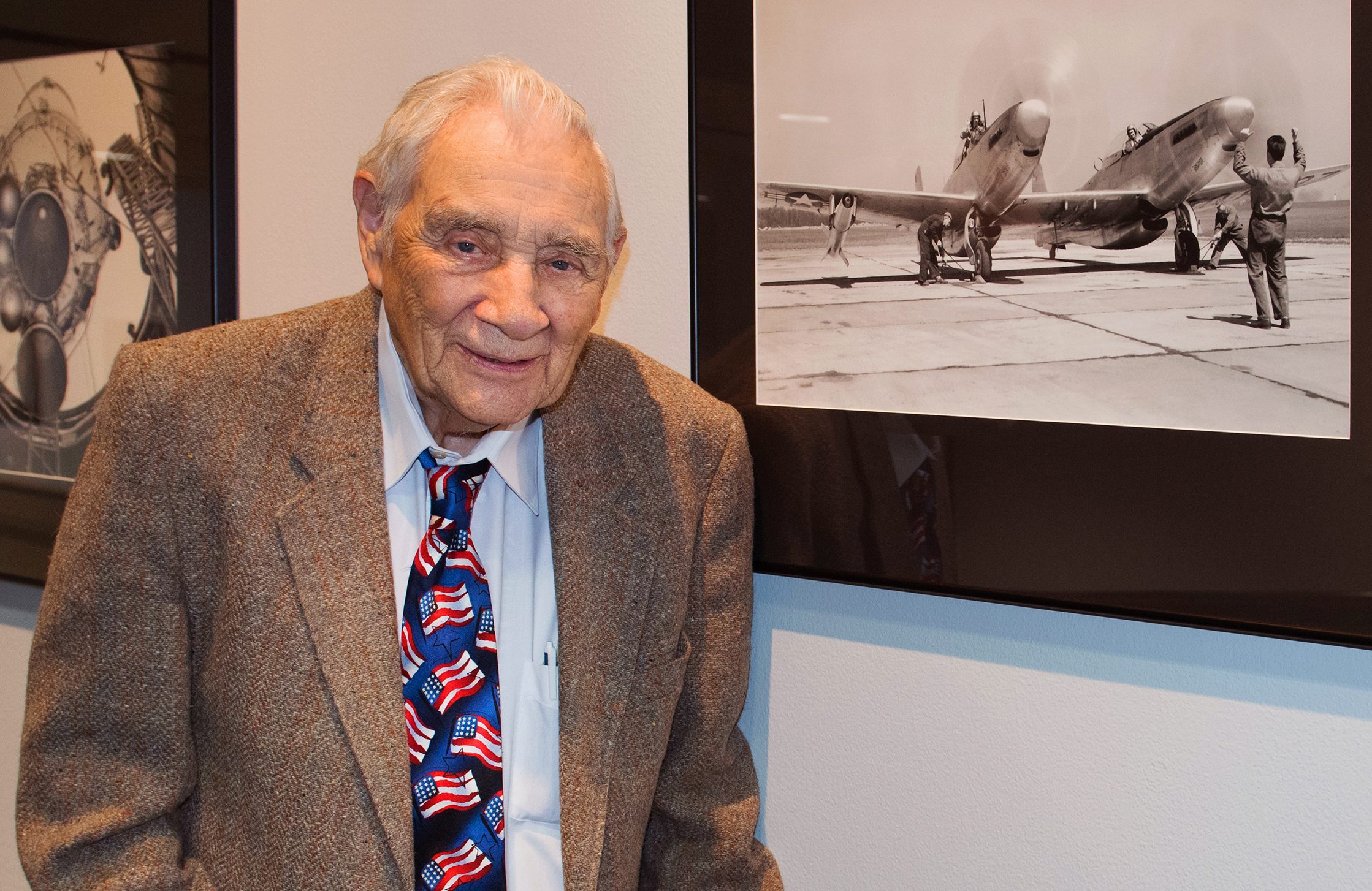 Elderly gentleman poses next to an old photograph displayed on a wall.