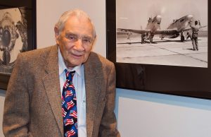 Elderly gentleman poses next to an old photograph displayed on a wall.